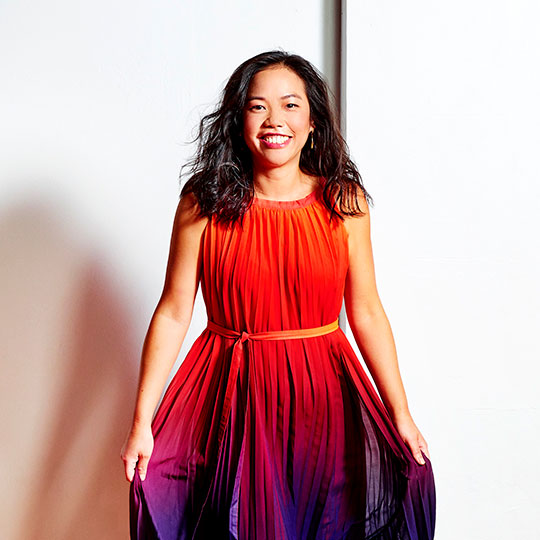 Andea Lam is looking directly at the camera and smiling, she has dark hair and is wearing a brighly cloured and pleated red, purple and blue dress, of which she is holding up the hem