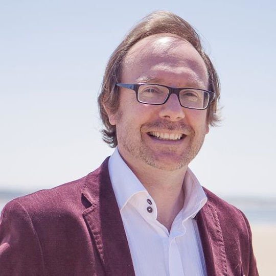 Matthew Hindson is pictured looking directly at the camera and smiling. He's wearing glasses and has short brown hair. Wearing a purple velvet jacket and light pink shirt.