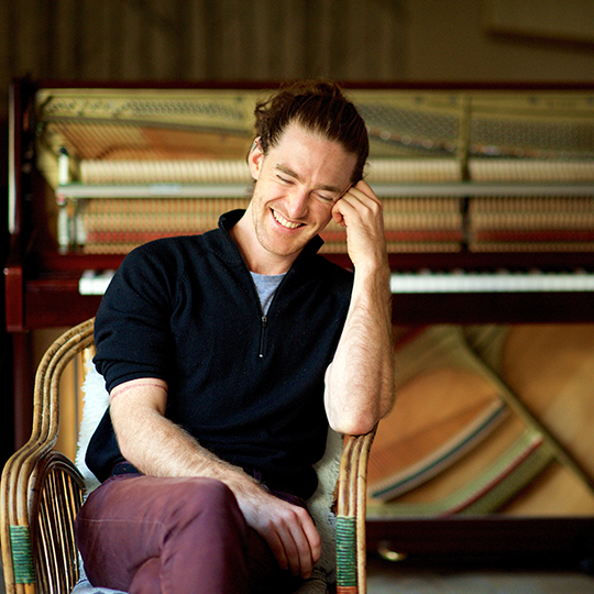 Robbie Mann, a young man with long hair tied up on his head, is sitting in a wicker chair in front of a piano, looking downward and laughing. He's wearing a blue top and purple jeans